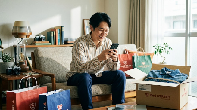 リユース品に囲まれたリビングで、スマートフォンを手に笑顔でセカンドストリートオンラインを見る男性。